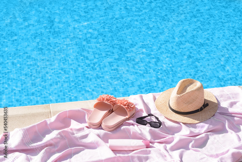 Pink blanket with slippers, hat, sunglasses and sunscreen near outdoor swimming pool on sunny day, space for text