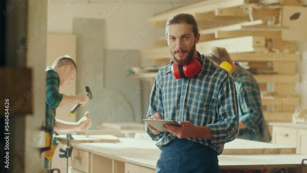 Portrait of Attractive Carpenter in Checkered Shirt and Construction Noise Cancelling Headphones on Neck Using a Tablet in a Loft Studio. Woodworking. Manual Labor. Carpentry, Handwork Concept. Portrait of Attractive Carpenter in Checkered Shirt and Construction Noise Cancelling Headphones on Neck Using a Tablet in a Loft Studio. Woodworking. Manual Labor. Carpentry, Handwork Concept.