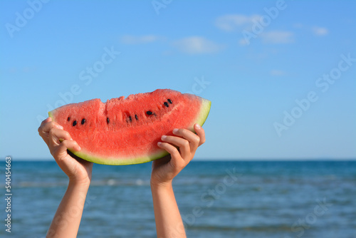 Child holding slice of fresh juicy watermelon near sea, closeup. Space for text