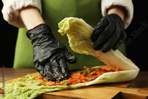 Woman preparing spicy cabbage kimchi at wooden table against dark background, closeup photo