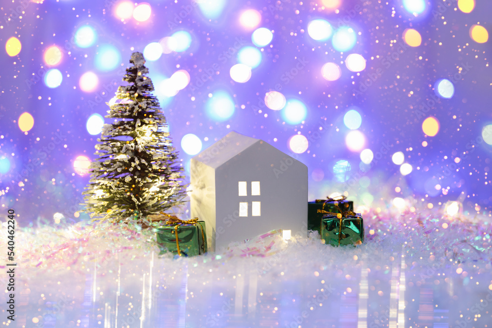 Christmas cute little house with tree in snow at night. Glowing festive garland on violet background with bokeh lights. Cozy atmosphere with home decor with selective focus. New Year card