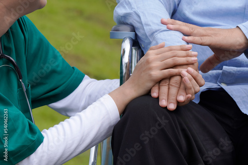 A young nurse gently holds the hand of an elderly man on a wheelchair with love and care.
