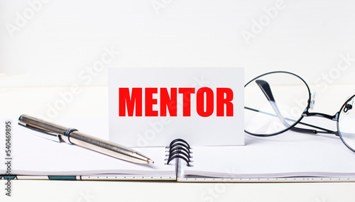 Close-up of a white card with the text MENTOR, a stylish pen and glasses on a desktop.