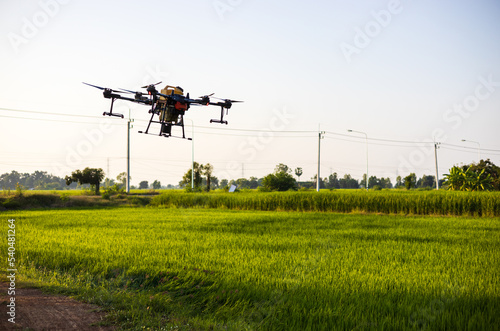 A view of a large drone in flight reflecting the midday sunlight to spray pesticides over the green paddy fields.