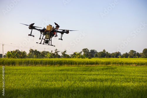 A view of a large drone in flight reflecting the midday sunlight to spray pesticides over the green paddy fields.