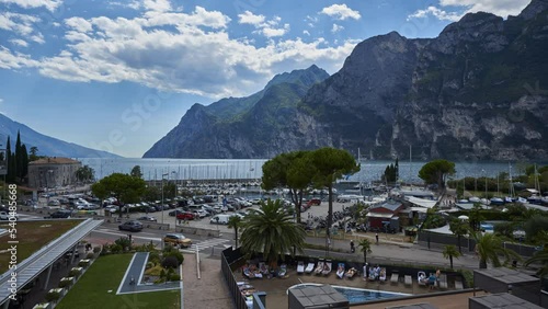 TimeLapse of the port area of Riva del Garda and the clouds moving over the lake and the mountains. Transport of vehicles along the lakefront and tourists who enjoy the services of the place.