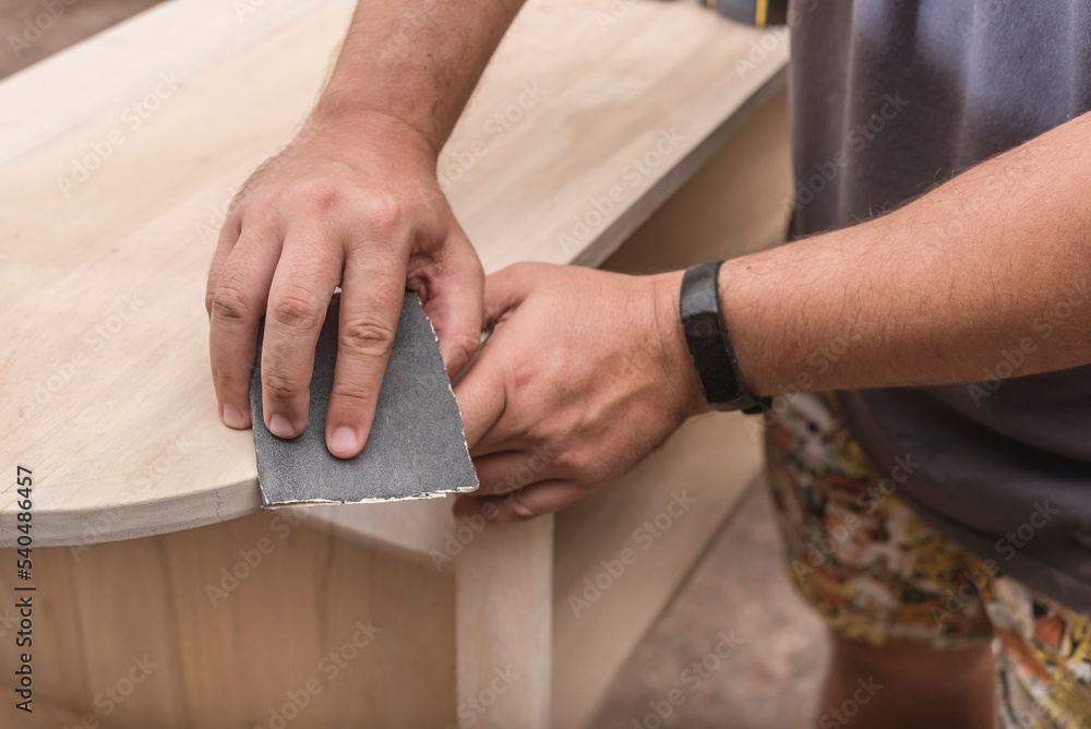 A man uses a piece of sandpaper to smooth out the edge of a circular table. Hand  sanding a wood surface. A DIY project at a personal workshop. Stock Photo |  Adobe