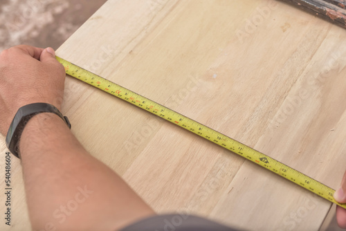 A man uses a tape measure to verify the width of a slab of wood to be used for furniture making.