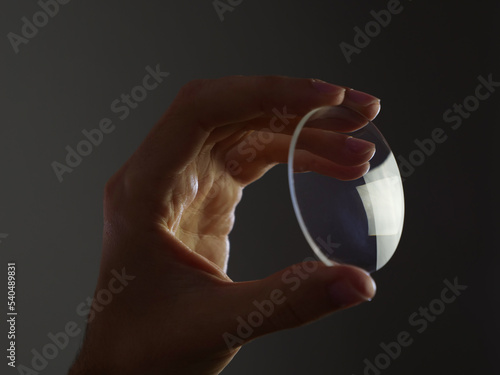 Hand holds an eyewear glass lens, glass prescription lens on dark background