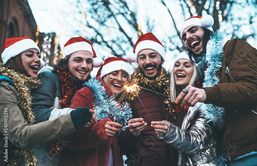 Happy friends wearing santa claus hat celebrating Christmas day holding sparklers - Cheerful young people having new year party outside - Winter traditional holidays concept