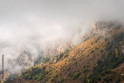 Mountains in a dense fog and sunny slope. Mystical landscape with beautiful sharp rocks in low clouds. Beautiful mountain foggy scenery on abyss edge with sharp stones.