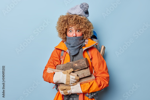 Surprised curly haired female camper prepares to make fire to warm herself carries wood wears knitted hat orange jacket and gloves rests in nature during severe cold winnter day isolated on blue wall photo