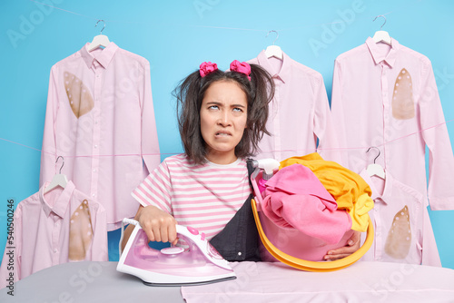 Displeased Asian woman has much work to do holds baket of laundry and electric iron stands near board wears striped t shirt poses in laundry room against blueblue background. Domestic chores photo