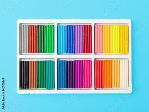 24 colors set of colorful plasticine sticks in white plastic packaging isolated on blue background. Pieces of multicolor plasticine modeling clay. Top view, closeup.
