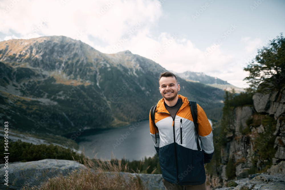 Naklejka premium Happy tourist on the top of the mountain against beautiful lake and forests on the hills. Concept of easy hike during the weekend. Hiker smiles and looks at the camera.