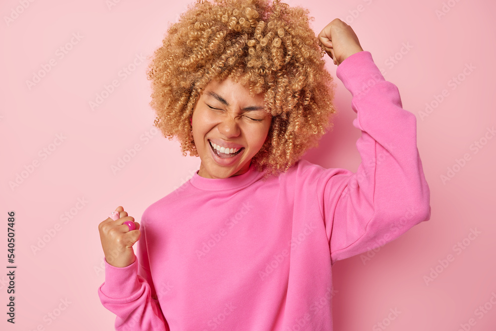 Optimistic curly haired young woman clenches fists makes triumph gesture celebrates success exclaims loudly wears casual jumper isolated over rosy background. People joy and celebration concept