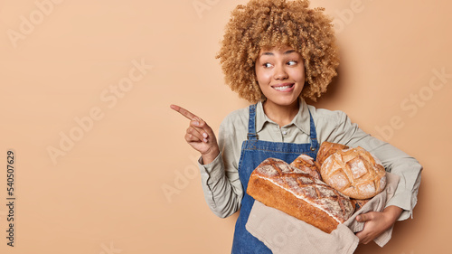Happy female baker wears shirt and denim sarafan works in bakery store holds freshly baked bread on linen towel bites lips points index finger aside demonstrates blank space for your advertisement