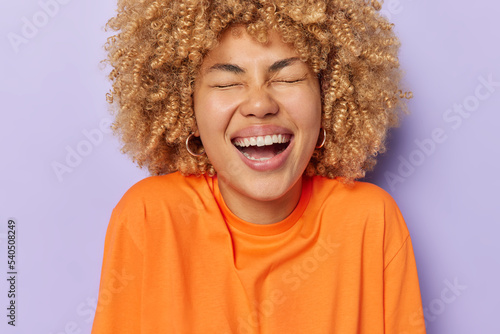 Cheerful young woman with curly bushy hair laughs happily keeps eyes closed expresses positive emotions feels very glad hears something funny wears orange jumper isolated over purple background