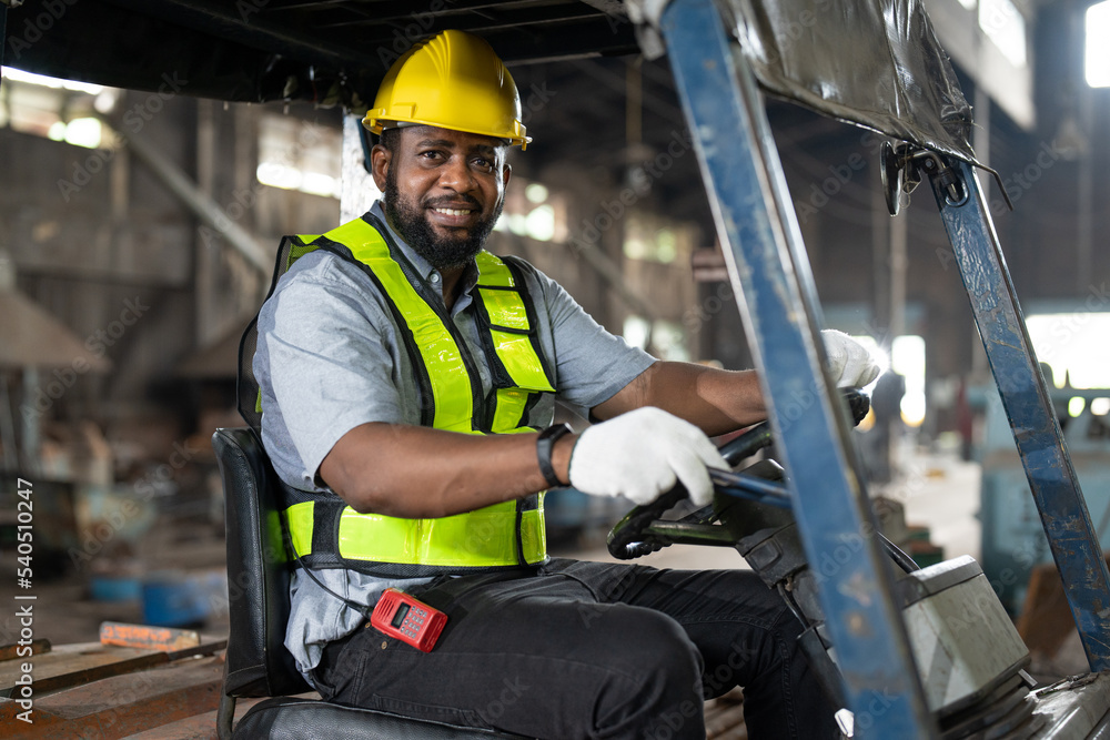 African American male worker driving forklift truck in heavy metal industrial factory. Smiling man engineer wearing vest and helmet safety working and moving heavy metal material at warehouse factory. African American male worker driving forklift truck in heavy metal industrial factory. Smiling man engineer wearing vest and helmet safety working and moving heavy metal material at warehouse factory.