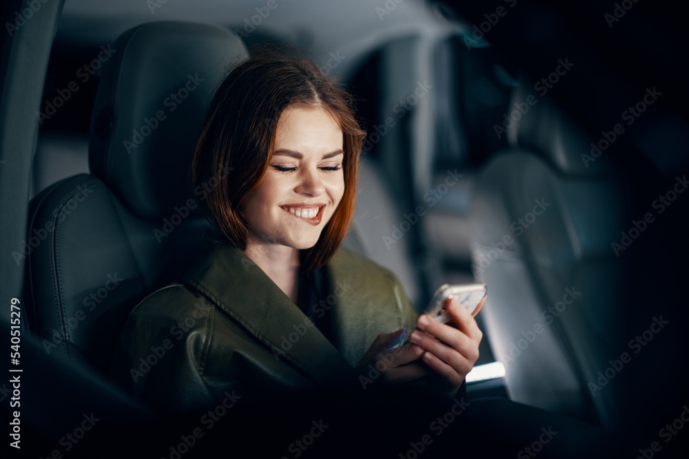 horizontal portrait of a stylish, luxurious woman in a green leather coat, sitting in a black car at night in the passenger seat, playfully biting her lower lip, holding her phone during the trip