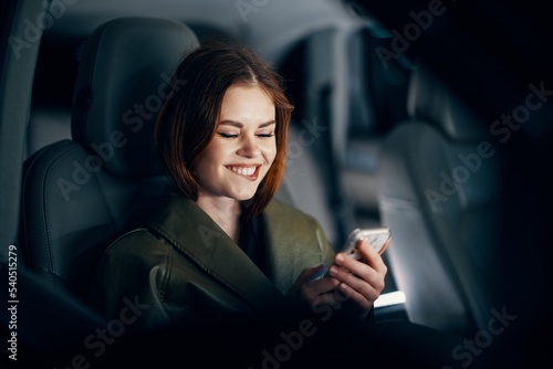 horizontal portrait of a stylish, luxurious woman in a green leather coat, sitting in a black car at night in the passenger seat, playfully biting her lower lip, holding her phone during the trip © SHOTPRIME STUDIO