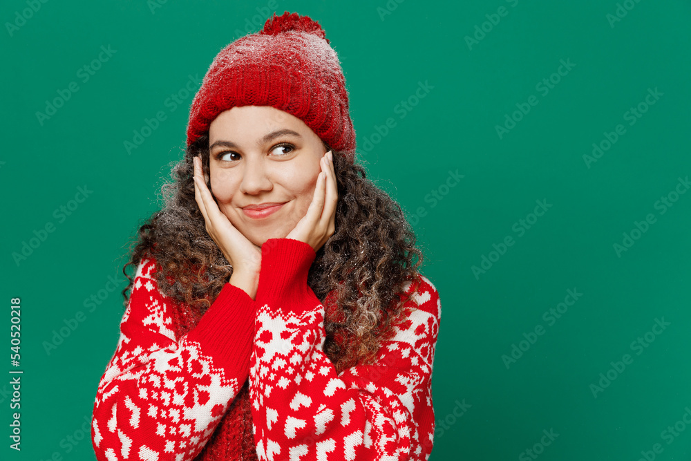 Merry young woman wearing red warm cozy knitted sweater hat posing hold face in reverie mood look aside isolated on plain dark green background studio. Happy New Year 2023 celebration holiday concept. Merry young woman wearing red warm cozy knitted sweater hat posing hold face in reverie mood look aside isolated on plain dark green background studio. Happy New Year 2023 celebration holiday concept.