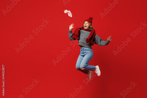Full body young excited woman wear grey sweater scarf hat jump high throw away napkin isolated on plain red background studio portrait Healthy lifestyle ill sick disease treatment cold season concept