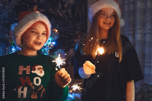 Happy teen girl wearing Santa hat, cute boy holding burning sparkler lights at Christmas tree background. New Year eve, Christmas celebration at home concept. Winter Holiday cheer. Festival Greeting © Natallia