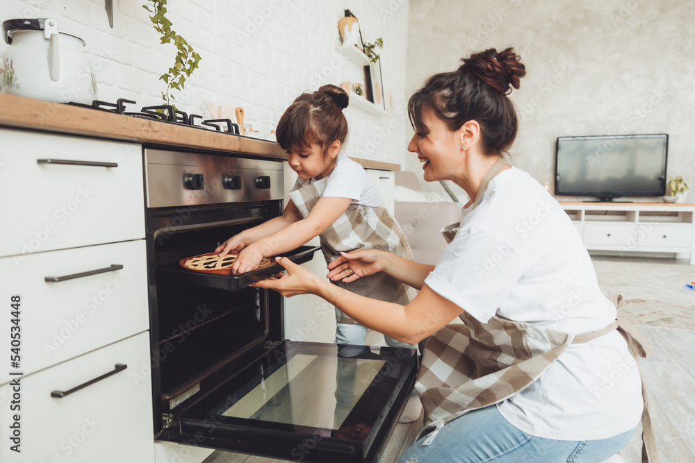 A little girl and her mom put a baking dish with a pie in the oven in the kitchen. mom and child cook a pie together in the kitchen. - obrazy, fototapety, plakaty 