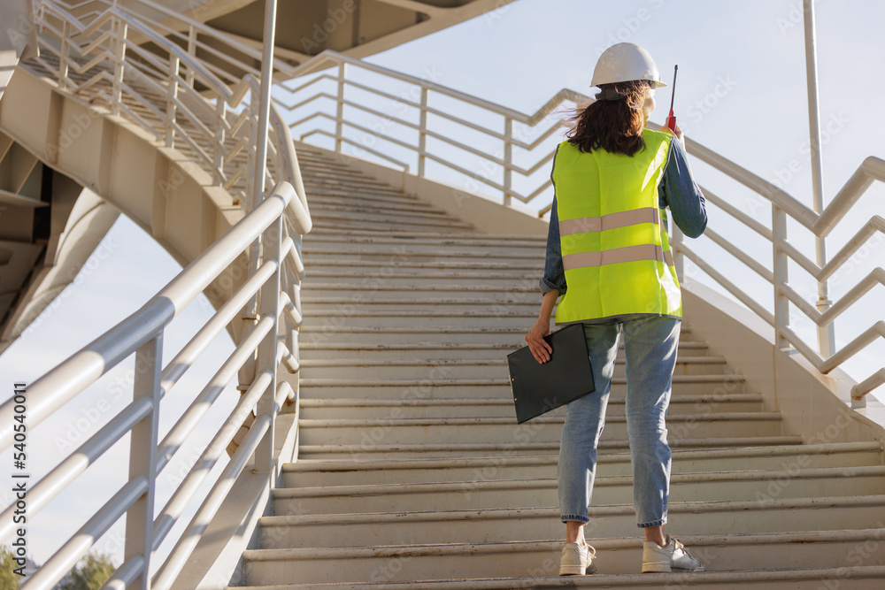 female engineer with a walkie-talkie at the construction site gives instructions. European successful female technologist or fire safety engineer works. foreman in a white helmet and protective yellow