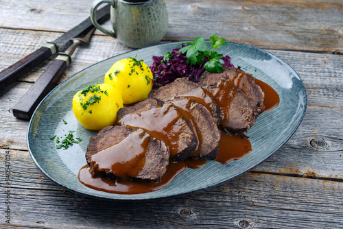 Traditionell geschmorter Sauerbraten vom Rind mit Blaukraut und Kartoffelklößen in würziger Dunkelbiersoße serviert als close-up auf einem Nordic design Teller