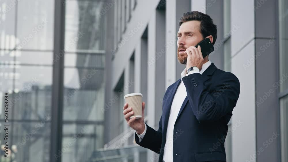 Young man standing outdoors talking on mobile phone holds paper cup with coffee professional businessman answering business call communicate with client male customer orders delivery using smartphone Young man standing outdoors talking on mobile phone holds paper cup with coffee professional businessman answering business call communicate with client male customer orders delivery using smartphone