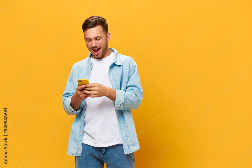 Happy smiling tanned handsome man in blue basic t-shirt type message chats with friends using phone posing isolated on yellow studio background. Copy space Banner Mockup. Online Social media concept Happy smiling tanned handsome man in blue basic t-shirt type message chats with friends using phone posing isolated on yellow studio background. Copy space Banner Mockup. Online Social media concept