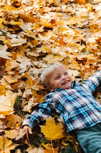 Happy little boy in yellow bright coat throwing leaves in sunny autumn forest and jumping joyfully