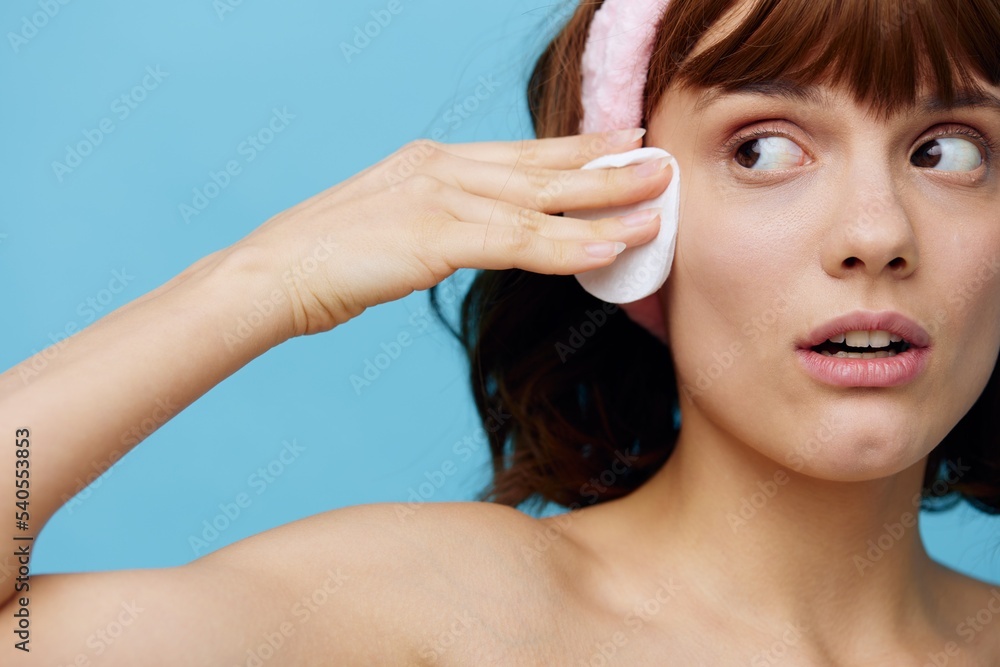 a woman with clean skin stands on a blue background in a white towel with a pink bandage on her head after a bath, removes makeup from her face with a cotton pad. Horizontal studio photography. a woman with clean skin stands on a blue background in a white towel with a pink bandage on her head after a bath, removes makeup from her face with a cotton pad. Horizontal studio photography.