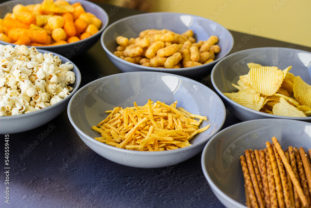 Salty snacks assortment shot on dark slate table. The picture includes potato chips, popcorn, peanut, cheese sticks, ketchup sticks, tzatziki chips, smoky flips, and others Salty snacks assortment shot on dark slate table. The picture includes potato chips, popcorn, peanut, cheese sticks, ketchup sticks, tzatziki chips, smoky flips, and others