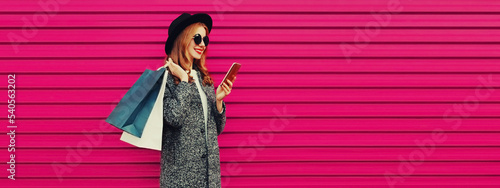 Portrait of happy smiling woman with shopping bags and smartphone wearing gray coat, hat on colorful pink background © rohappy