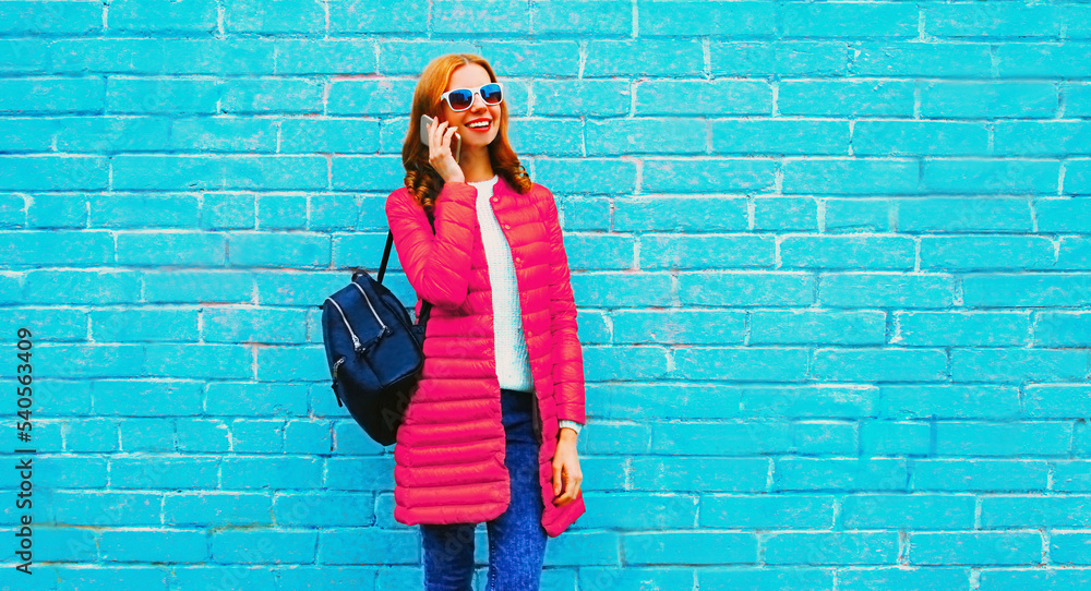 Portrait of happy smiling young woman calling on smartphone wearing pink jacket, backpack on blue background Portrait of happy smiling young woman calling on smartphone wearing pink jacket, backpack on blue background