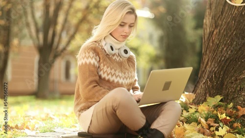 Beautiful blonde woman have distance work or study online sitting in the yellow autuwn park. Smiling young woman with headphones on her neck typing on laptop. photo