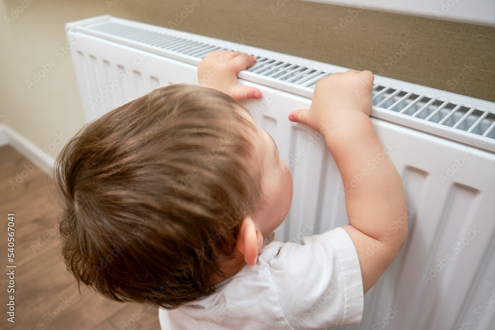 Toddler baby holding on to the radiator, child hand on the heating system close-up. White radiator for heating the home room and children's hand Toddler baby holding on to the radiator, child hand on the heating system close-up. White radiator for heating the home room and children's hand