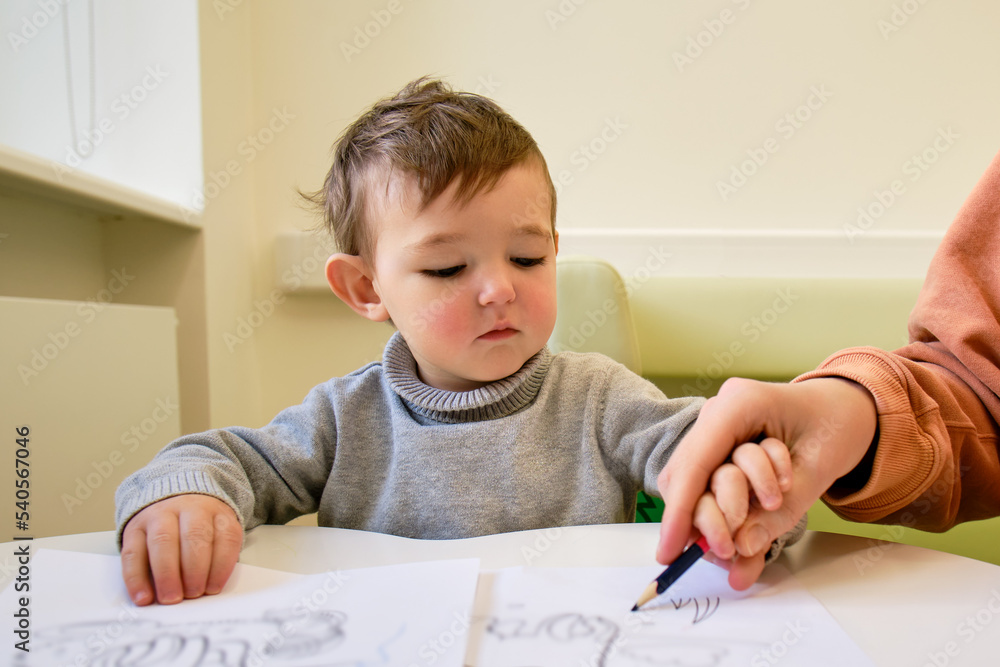 Mother teaches to draw toddler baby with a pencil on a white sheet of paper. Parent with child boy learning to color coloring book. Kid aged one year and three months Mother teaches to draw toddler baby with a pencil on a white sheet of paper. Parent with child boy learning to color coloring book. Kid aged one year and three months
