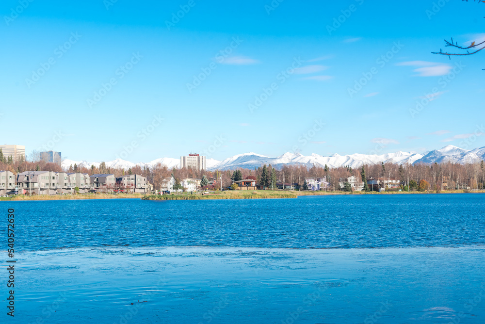 Westchester Lagoon with small island on the lake and row of duplex houses with downtown Anchorage buildings office towers in background