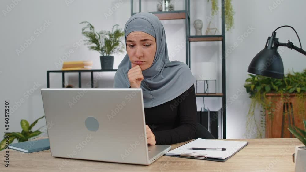Business woman in hijab thinking about an important creative project work decision at home office desk. Confident muslim freelancer girl busy occupation looking through analytical reports on laptop Business woman in hijab thinking about an important creative project work decision at home office desk. Confident muslim freelancer girl busy occupation looking through analytical reports on laptop