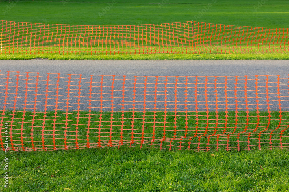 Fototapeta premium Plastic orange barricade net along sidewalk with green grass, Construction safety fence protected from construction site work, Closed area for maintenance or repair under ground of street in the park.