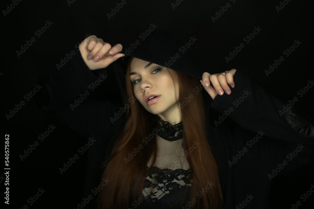 portrait of a beautiful young red-haired girl on a dark black background in the studio, with a cleavage and big breasts in black hood, hoodie portrait of a beautiful young red-haired girl on a dark black background in the studio, with a cleavage and big breasts in black hood, hoodie