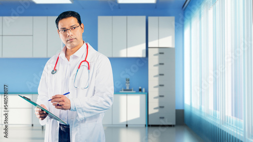 Doctor therapist. Medical worker in white coat. Portrait therapist taking notes. Man is standing indoors in clinic. Blurred hospital interior. Therapist is holding clipboard. Doctor with stethoscope