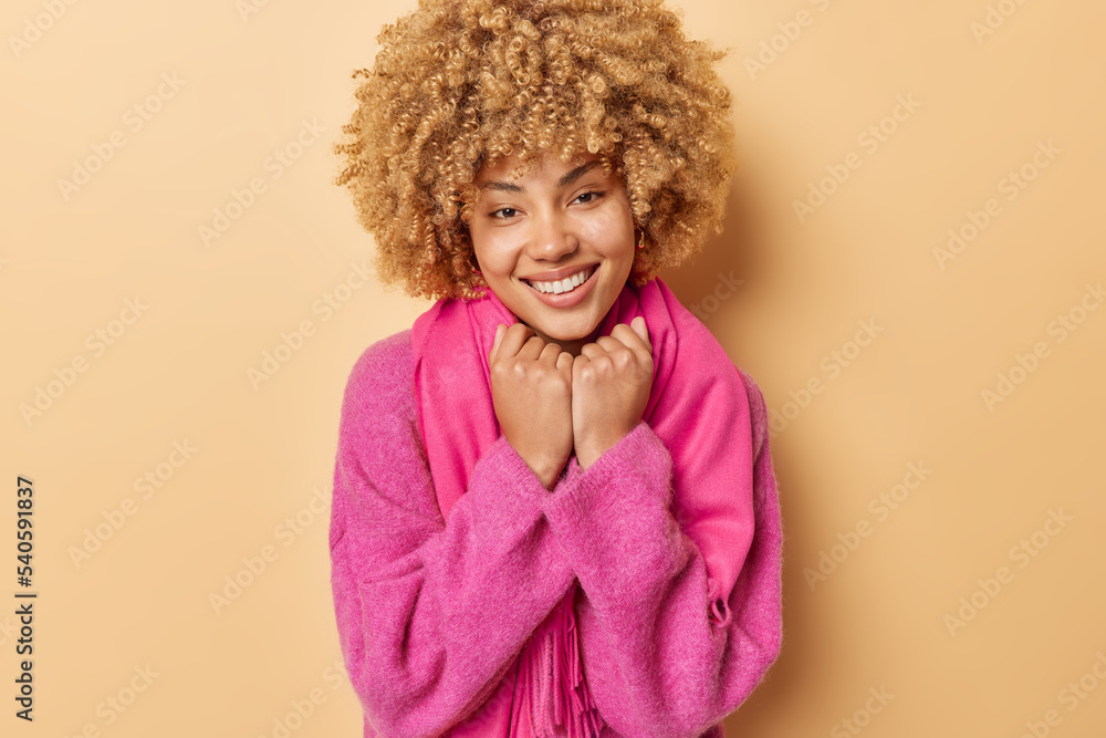 Portrait of good looking woman with curly hair keeps hands under chin smiles toothily wears pink jumper and scarf isolated over beige background. Positive emotions and genuine happiness concept Portrait of good looking woman with curly hair keeps hands under chin smiles toothily wears pink jumper and scarf isolated over beige background. Positive emotions and genuine happiness concept