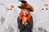 Mysterious woman has long orange hair keeps hands together wears hat decorated with autumn leaves and black silk dress prepares for Halloween celebration made creepy ghosts for decorating room
