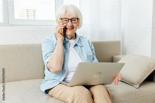 a happy, pleasant elderly lady with white hair is sitting on a cozy sofa in a bright apartment holding a laptop on her lap, holding it with her hand, laughing loudly with happiness © Tatiana