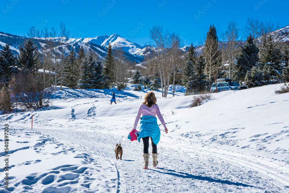 Woman walking her dog in snow on sunny  winter day;  mountains and blue sky in background